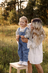 Blonde woman and little child standing on sunlit field. Mother in white dress, child in denim...