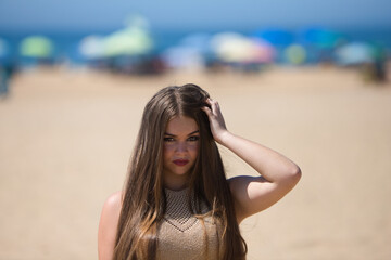 portrait of beautiful young blonde woman with long hair touches her hair with her hands while looking at the camera. The girl is on the beach. In the background the blue sea and umbrellas.