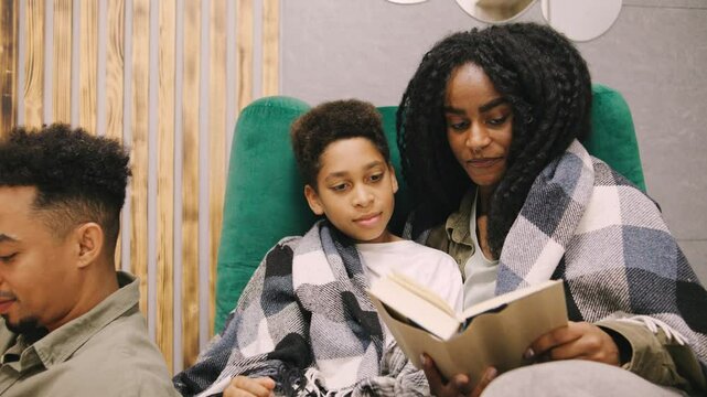 An African American mom and son enjoy reading a book together in a cozy chair, while dad works on his laptop nearby, blending family time with productivity.