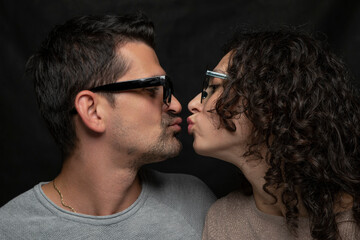 A young man and a young woman wearing sunglasses are about to kiss each other on the mouth against a black background