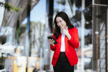 Beautiful Asian businesswoman walking confidently on a modern city street, smiling while using a smartphone. Success, urban lifestyle and modern communication.