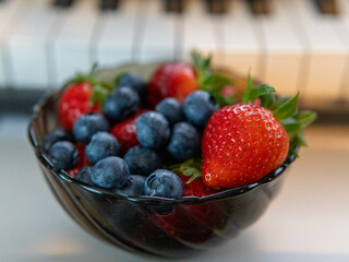 Close-up of fresh strawberries and blueberries in a black bowl with a blurred piano keyboard in the background. Vibrant colors emphasize the freshness and appeal of the fruit.
