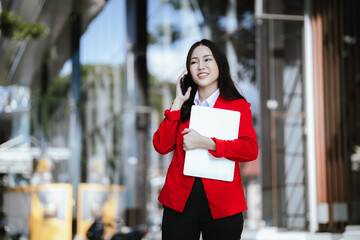 Using laptop, video call, Smiling young Asia business woman leader entrepreneur in red color suit working outdoor urban street walk.