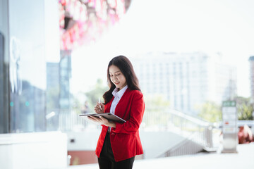Fototapeta premium Using laptop, video call, Smiling young Asia business woman leader entrepreneur in red color suit working outdoor urban street walk.