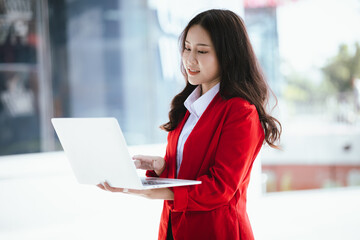 Using laptop, video call, Smiling young Asia business woman leader entrepreneur in red color suit working outdoor urban street walk.