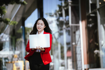 Using laptop, video call, Smiling young Asia business woman leader entrepreneur in red color suit working outdoor urban street walk.