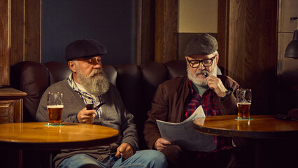 Two elderly bearded men in caps sitting in leather armchairs, smoking tobacco pipes, reading newspaper and drinking bee in cozy bar. Concept of elderly people lifestyle, drinks, retro fashion