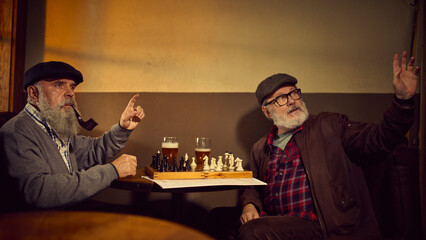 Two elderly bearded men in caps, sitting in cozy bar at wooden table with chess and ordering beer. Concept of elderly people lifestyle, drinks, retro fashion