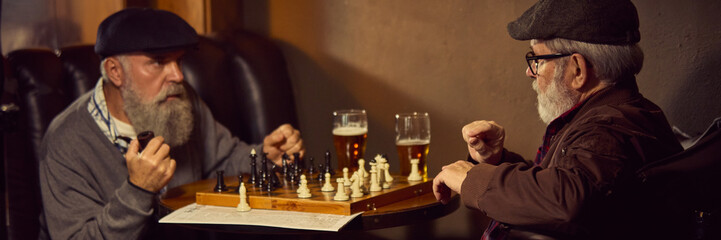 Two elderly men enjoying quiet game of chess and glasses of beer in cozy bar. Relaxed senior...
