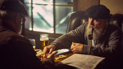 Elderly man with pipe in mouth playing dominoes with his friend in warm, cozy bar setting,drinking beer, quiet leisure moment. Concept of elderly people lifestyle, drinks, retro fashion