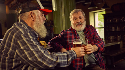 Two elderly men in checkered shirts sitting at modern cozy bar, drinking beer, involved in conversation, and enjoying sport translation. Concept of elderly people lifestyle, drinks, retro fashion