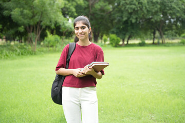 portrait of young indian college girl with backpack and notebooks standing and smiling in public park or garden.