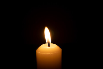 A single burning candle flame or light glowing on a big white candle isolated on black or dark background on table in church for Christmas, funeral or memorial service