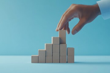 Hand stacking wooden blocks into a step formation with an arrow pointing upwards, representing the process of achieving business success, with a minimalist blue backdrop and space for text.