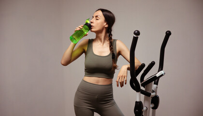 Woman in green activewear taking refreshing sip from water bottle while leaning on an elliptical...