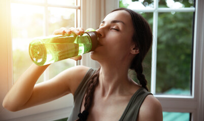 A young sporty woman with braided hair is drinking water from a green bottle, sitting by a window with natural light, creating a peaceful, refreshing atmosphere in a cozy indoor space.