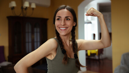 Smiling athlete young woman with braided hair flexes her arm confidently in a cozy indoor setting. The natural light highlights her positive energy and strength, capturing a moment of empowerment