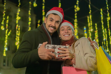 Smiling couple wearing Santa hats exchanging gifts during Christmas shopping night, standing under festive lights with gift boxes and shopping bags, enjoying holiday celebrations and seasonal joy