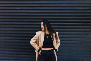 Stylish young asian woman posing with hands on hips in front of a metal shutter, looking away from...