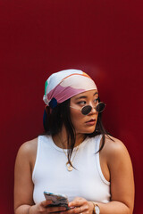 Fashionable young asian woman wearing a headscarf and sunglasses using a smartphone with a vibrant red background