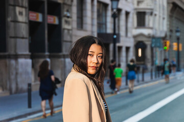 Fototapeta premium Stylish young asian woman posing confidently on a bustling city street, wearing a beige coat