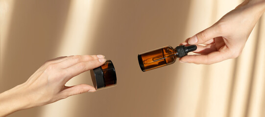 Two female hands. In one hand there is an amber bottle with serum, in other glass jar with...