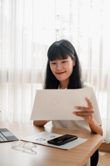 A young woman sits at a desk reviewing documents, surrounded by modern technology and natural light from a window.