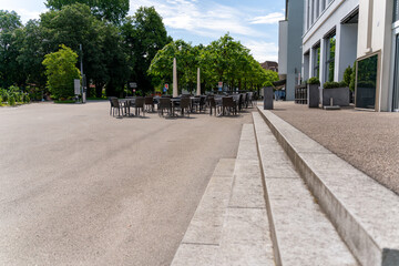 Naklejka premium Place in the city with empty chairs and tables at the outside in front of a cafe. Scene viewed from some distance. Beautiful day with blue, slightly clouded sky.