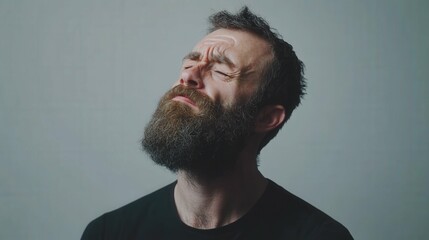 Obraz premium Bearded man with a grimace holding his jaw as if experiencing pain, standing against a plain background under soft studio lighting