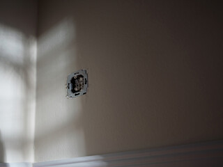 Single uninstalled wall socket with exposed wiring, casting shadows on a beige wall, suggesting electrical work and interior renovations