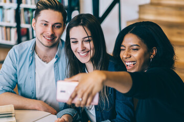 Laughing multiracial students taking selfie on smartphone at table in library