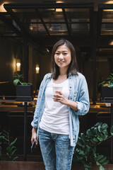 Cheerful adult pretty female surfing mobile while drinking takeaway coffee in front of cafe on street