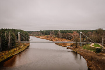 Aerial view of a suspension bridge crossing a calm river with surrounding autumnal trees, fields,...
