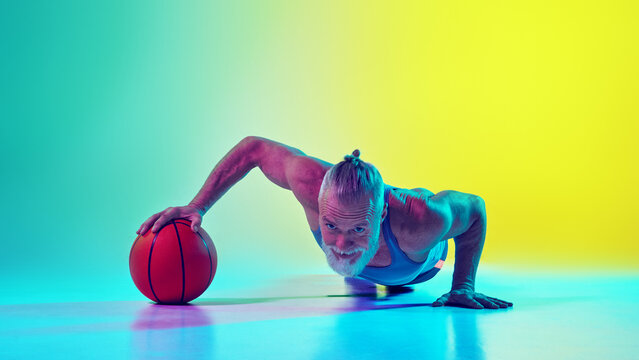 Muscular elderly man in sportswear doing push up exercise with one hand on basketball ball against gradient green yellow background in neon light. Concept of sport, active elderly lifestyle, health
