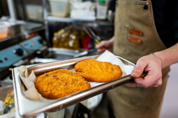 A person in an apron holds a metal tray with two large, golden-brown fried items, possibly schnitzels, in a commercial kitchen setting. The background shows kitchen equipment and utensils