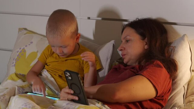 Child and mother lying in bed together in bedroom at home, child playing on tablet, woman helping boy to complete level in game. Boy talking.