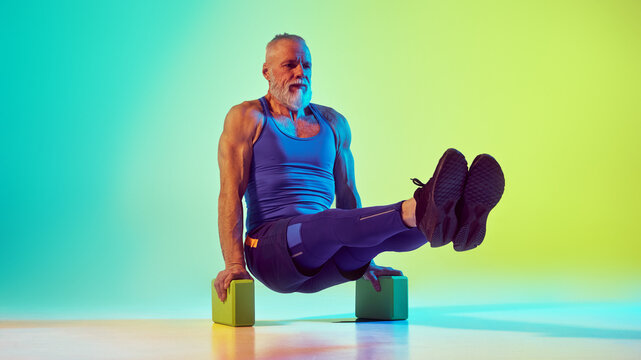 Sportive senior man in sportswear training, doing exercised with yoga blocks against gradient green yellow background in neon light. Concept of sport, active elderly lifestyle, health care