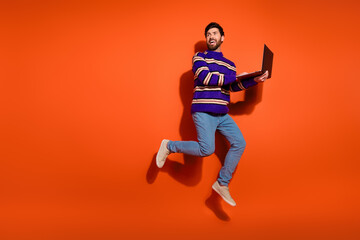 Full size photo of nice young male jump look empty space netbook wear trendy striped violet outfit isolated on orange color background