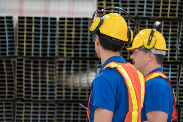 Two construction workers in reflective safety vests and helmets engage in factory industrial site. Both wear ear protection, emphasizing a noisy environment, with metal beams in the background.