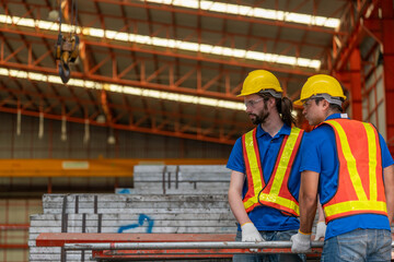 Two construction workers in reflective safety vests and helmets engage in conversation at industrial site. Both wear ear protection, emphasizing a noisy environment, with metal beams in the background