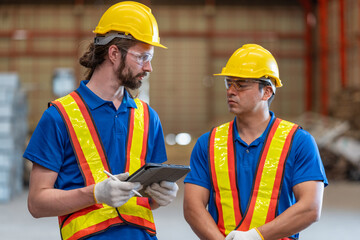 Two construction workers in reflective safety vests and helmets engage in conversation at industrial site. Both wear ear protection, emphasizing a noisy environment, with metal beams in the background