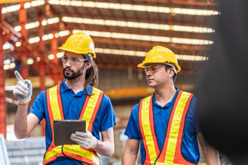 Two construction workers in reflective safety vests and helmets engage in conversation at industrial site. Both wear ear protection, emphasizing a noisy environment, with metal beams in the background