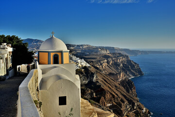Die Kirche Agios Stylionos auf der Griechischen Insel Santorin steht direkt an der Caldera bei der Stadt Fira. Man sieht schön der verlauf der steilen Abbruchkante der Caldera im warmen Abendlicht.