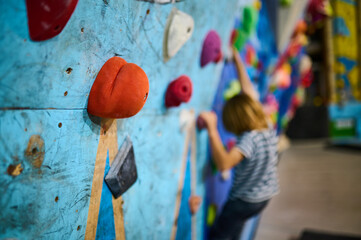 Close-up of climbing holds on a colorful indoor bouldering wall, with a young climber blurred in...