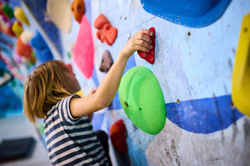 A 6 year old boy focused on climbing, carefully gripping small holds on an indoor bouldering wall. The colorful holds and detailed concentration showcase his determination © sandsun