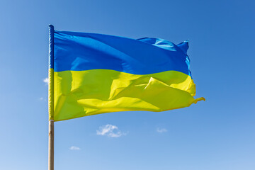 The state flag of Ukraine flutters in the wind. A yellow and blue flag on a wooden pole against a clear blue sky on a sunny day.