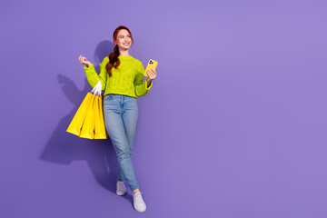 Cheerful young woman in bright green sweater holding shopping bags and smartphone on purple background.