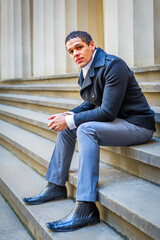 A man is sitting on concrete steps, wearing a black coat and gray trousers. He looks thoughtfully towards the camera, with his hands clasped together.