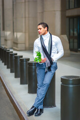 A person leans against a series of cylindrical posts on a city street, holding a sprig of greenery. Dressed in a white shirt, grey trousers, and a black scarf, they appear thoughtful.