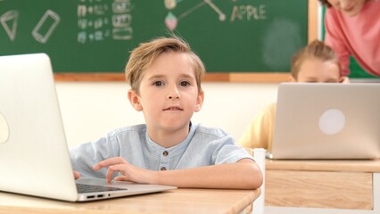 Attractive caucasian boy looking at camera while studying math at school. Diverse cute student doing test while teacher asking asian child question and point at paper in front of blackboard. Pedagogy.